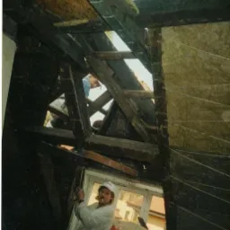 Pracownik w czapce z papierosem w ustach, w trakcie prac związanych z izolacją poddasza, widoczne drewniane elementy konstrukcyjne dachu i częściowo ułożona wełna mineralna, pakunki wełny...